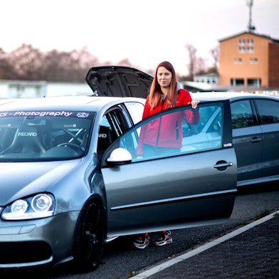 Michelle Engel steht an ihrem Golf R32. Im Hintergrund schauen zwei Männer auf ihr Handy.