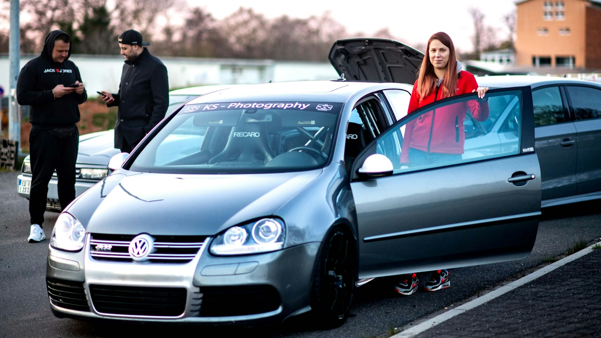 Michelle Engel steht an ihrem Golf R32. Im Hintergrund schauen zwei Männer auf ihr Handy.