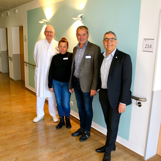 Drei Männer und eine Frau posieren lächelnd für eine Bildaufnahme vor einer Wand im Krankenhaus. Links im Hintergrund sind Türen zu sehen, die zu Patientenzimmern führen. Rechts oben im Bild ist ein Schild mit der Aufschrift "214" zu sehen.