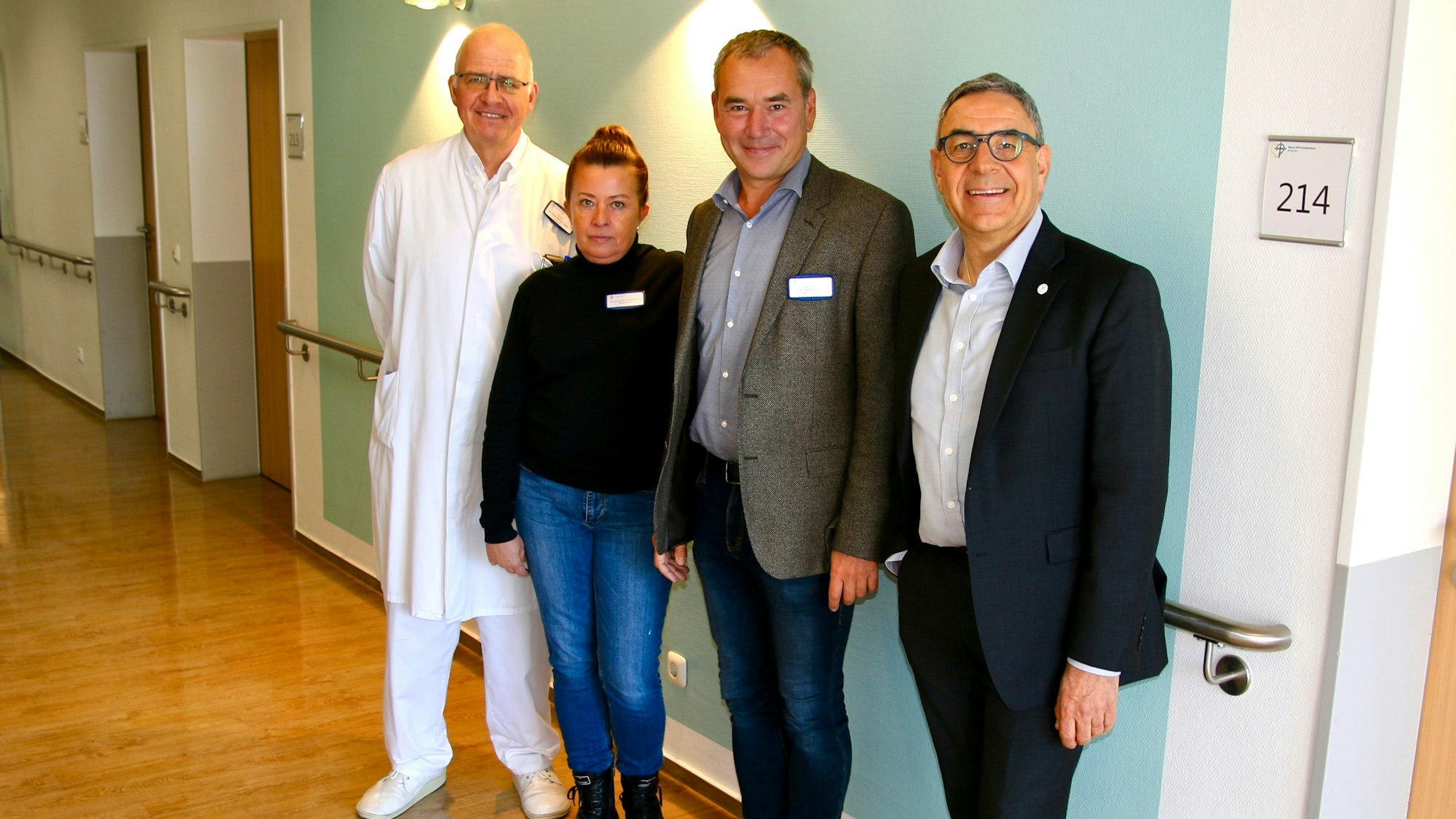 Drei Männer und eine Frau posieren lächelnd für eine Bildaufnahme vor einer Wand im Krankenhaus. Links im Hintergrund sind Türen zu sehen, die zu Patientenzimmern führen. Rechts oben im Bild ist ein Schild mit der Aufschrift "214" zu sehen.