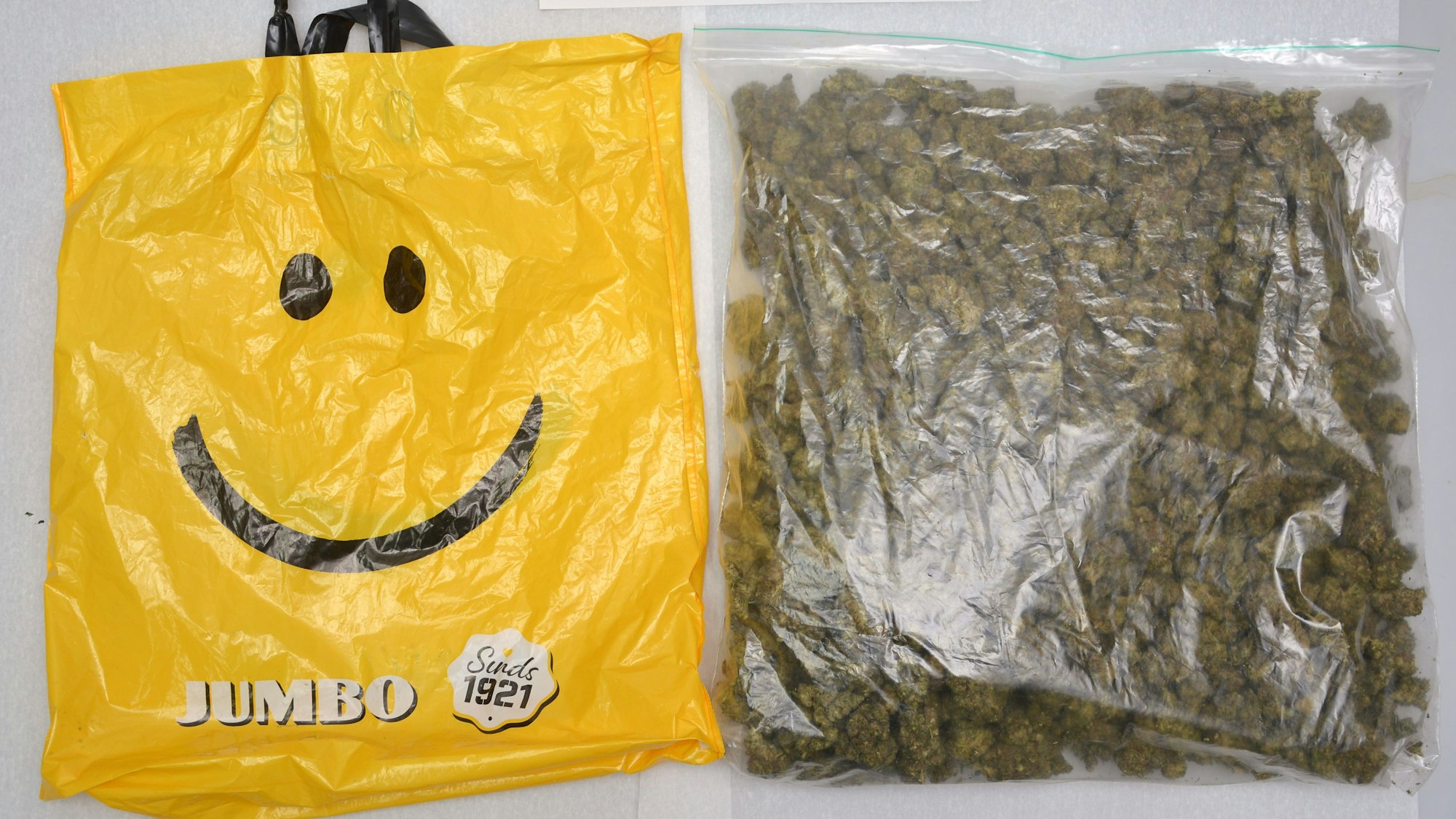 Eine gelbe Plastiktüte, auf der ein Smiley zu sehen ist. Darin transportierte der Mann ein Kilogramm Marihuana, das daneben zu sehen ist.