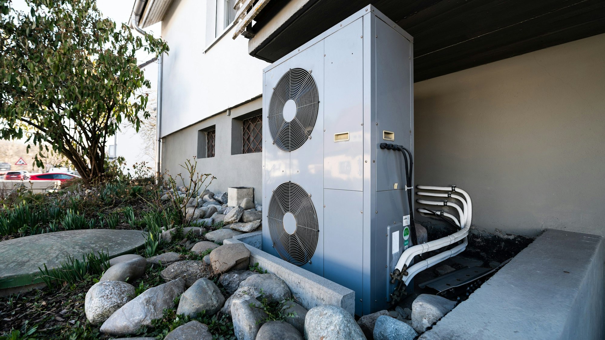 Im Vorgarten eines zweigeschossigen Hauses steht eng vor der Hauswand und unter einem Balkon die Lüftungsanlage einer Wärmepumpe.