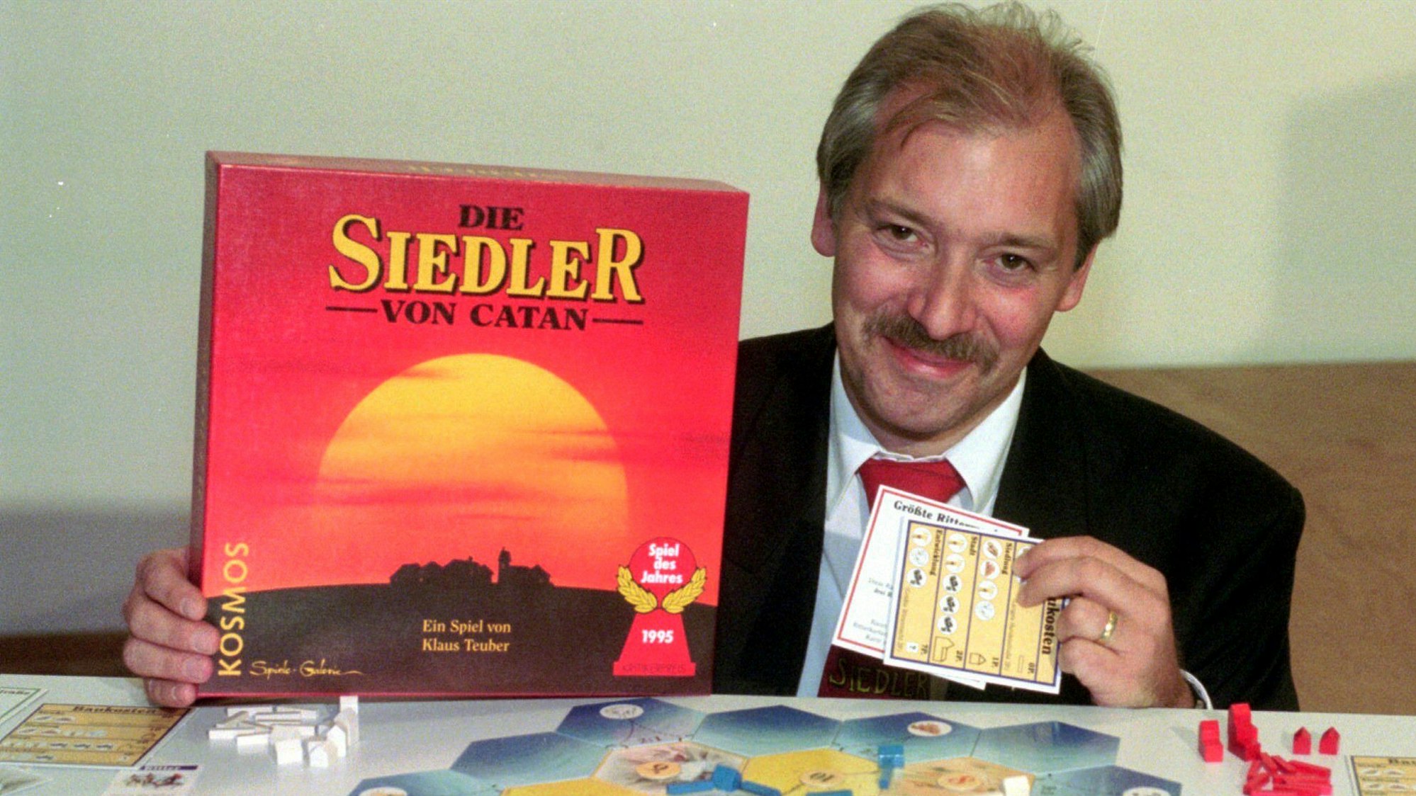 Klaus Teuber sitzt an einem Tisch, in der Hand hält der die Spielschachtel von „Catan“, vor ihm ist das Spiel aufgebaut.