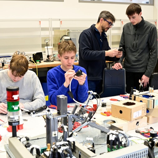 In einer Werkstatt sind zwei Azubis an Steckern beschäftigt. Im Hintergrund erklärt der Ausbilder einem weiteren Azubi etwas.