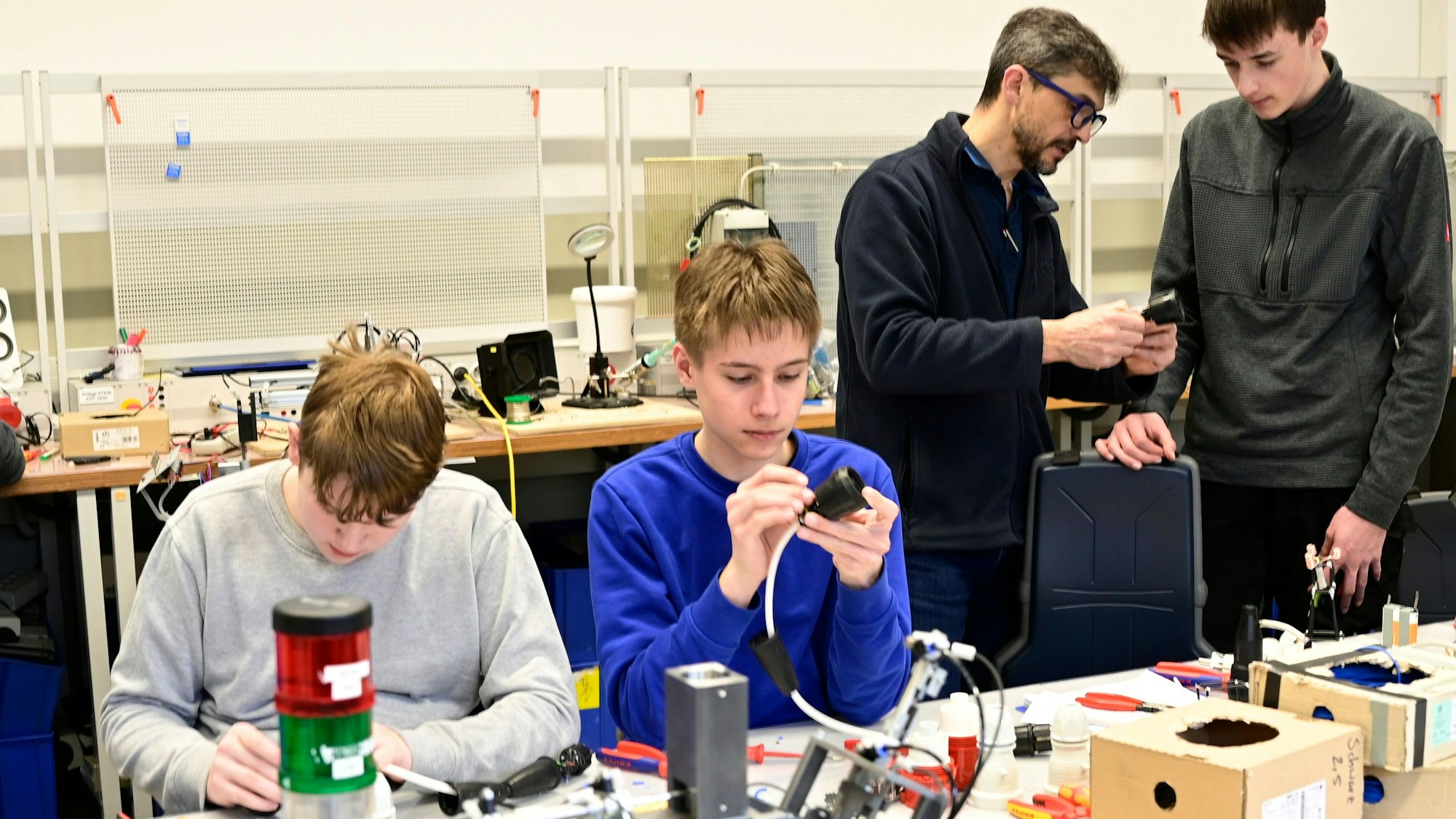 In einer Werkstatt sind zwei Azubis an Steckern beschäftigt. Im Hintergrund erklärt der Ausbilder einem weiteren Azubi etwas.