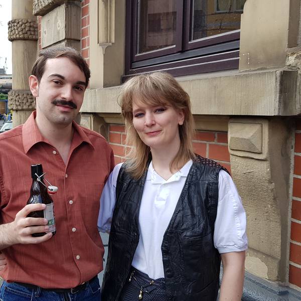 Die beiden sind gekleidet wie in den 60er-Jahren. Luca trägt einen Schnurbart und hält ein Bier in der Hand. Sabine trägt ein weißes Top mit einer schwarzen Weste.