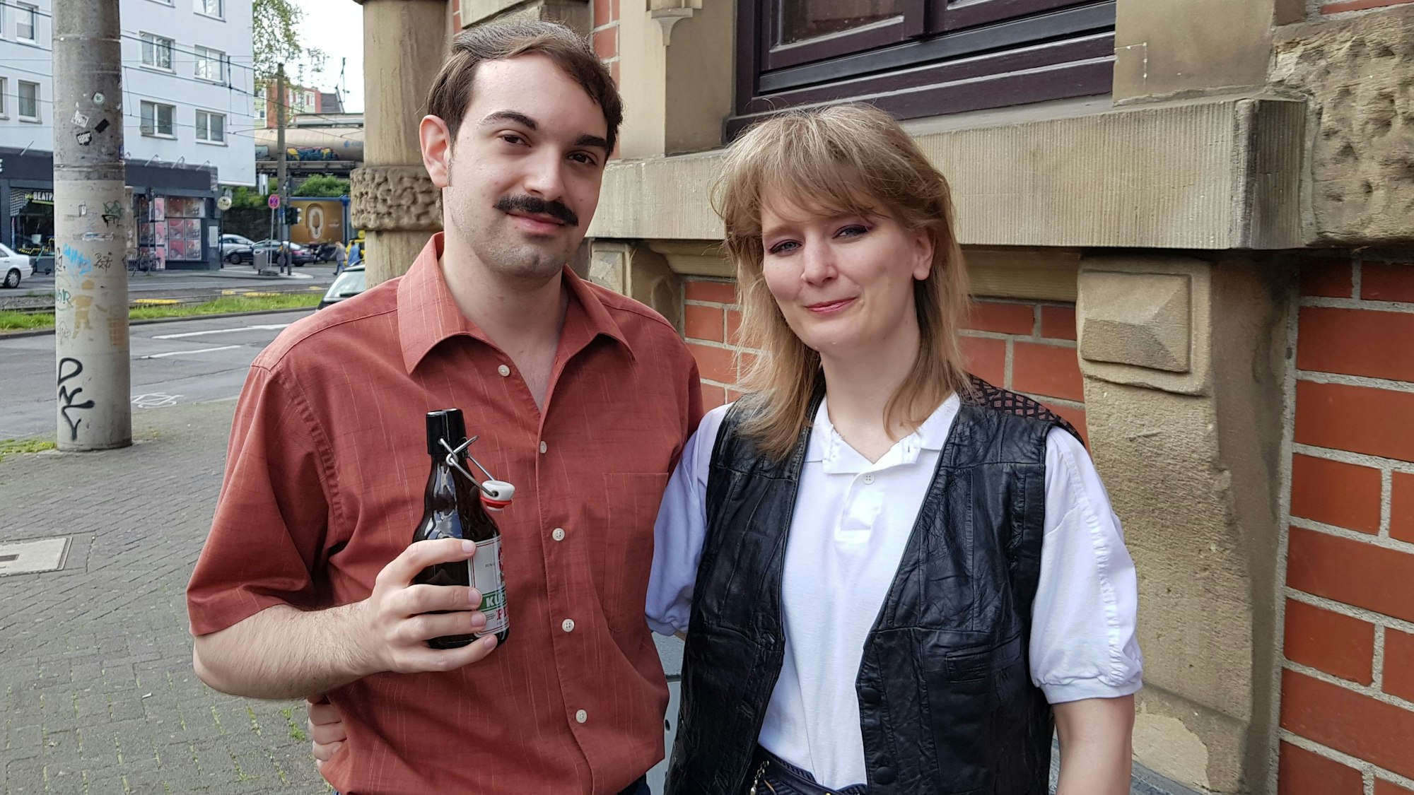Die beiden sind gekleidet wie in den 60er-Jahren. Luca trägt einen Schnurbart und hält ein Bier in der Hand. Sabine trägt ein weißes Top mit einer schwarzen Weste.