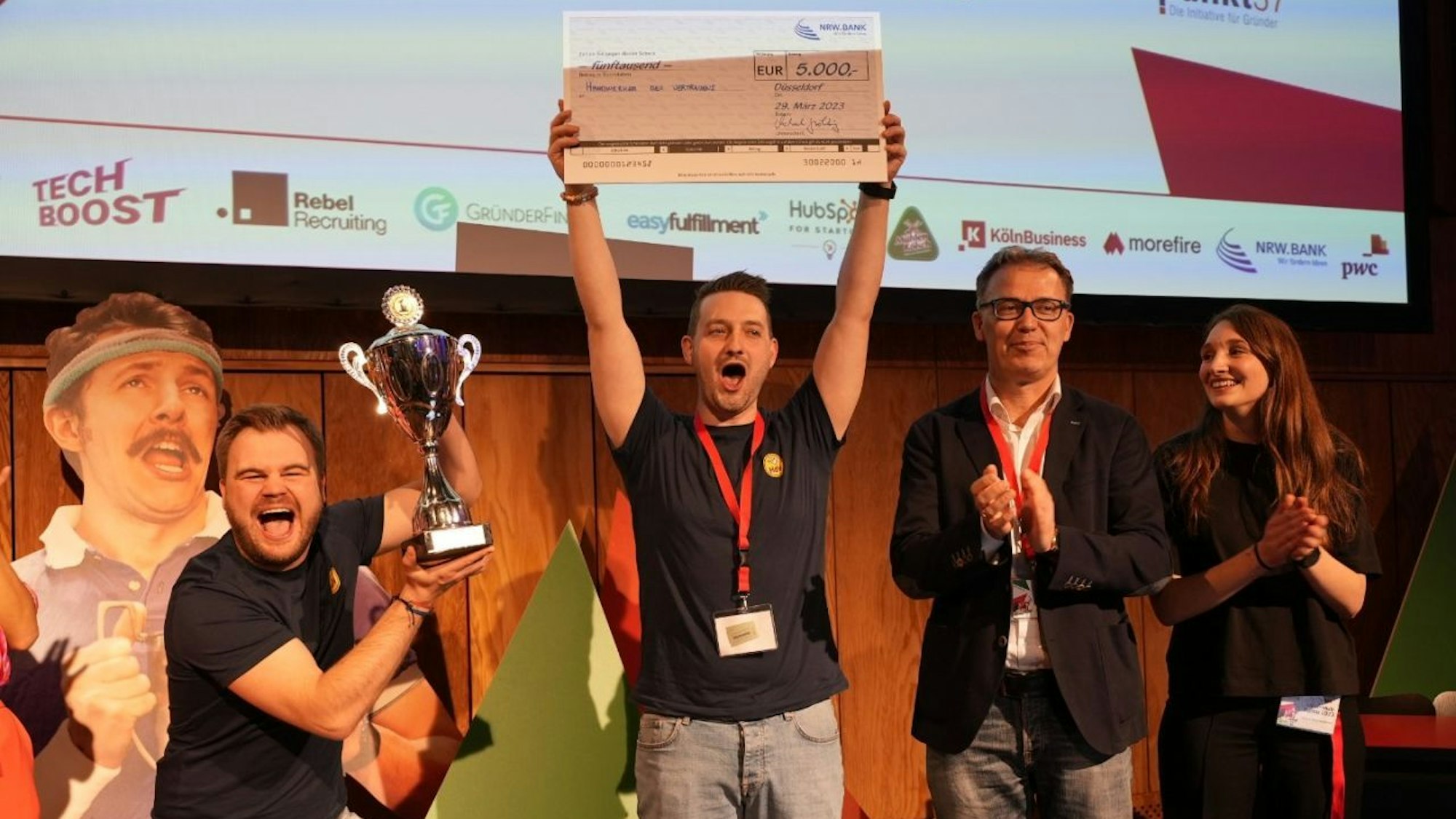 Mathias (l.) hält den Pokal in den Händen, Daniel zeigt den Check. Daneben applaudieren ein Mann und eine Frau.