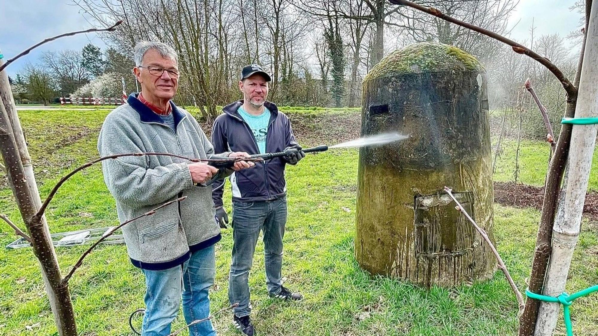 Die beiden Männer stehen seitlich vor dem Bunker und säubern ihn mit einem Hochdruckreiniger.