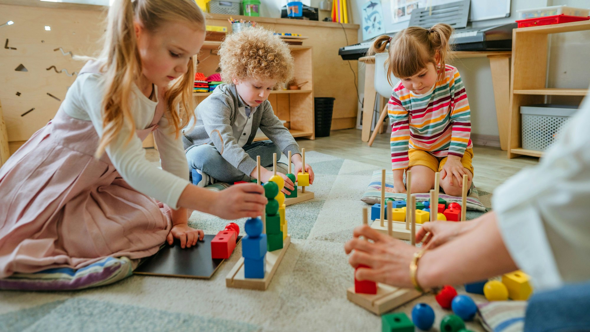 Drei Kinder sitzen im Innenraum eines Kindergarten auf dem Boden und spielen mit Bauklötzen.