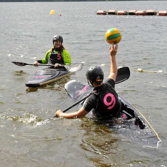 Zwei Kanupolo-Spielende der Schülerklasse sitzen rudernd in ihren Einer-Kanus. Der vorne im Bild befindliche Kanupolospieler wirft einen Ball zu einer Kanupolospielerin. Um die beiden herum ist Wasser.