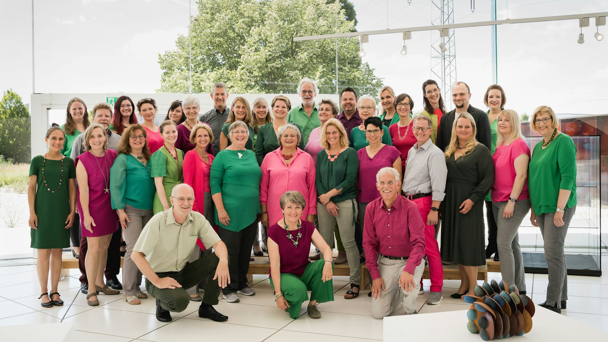 Der Chor posiert als Gruppe. Die Teilnehmer tragen rosa-pinke und grüne Kleidung.