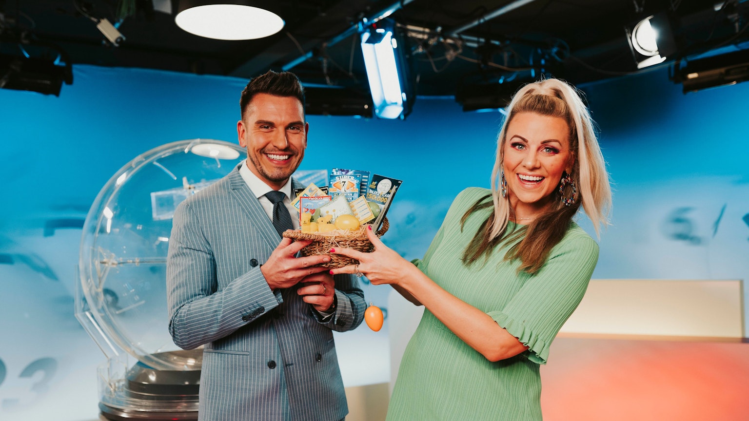 Die Lottomoderatoren Chris Fleischhauer und Miriam Hannah stehen im Lotto-Studio und halten ein Körbchen mit Rubbellosen in den Händen.