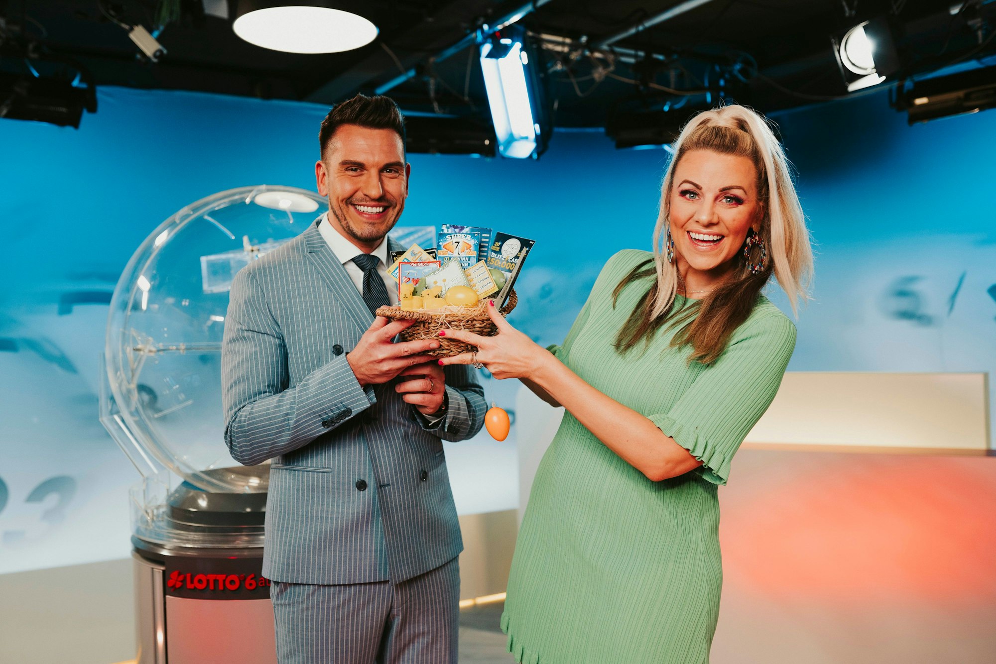 Die Lottomoderatoren Chris Fleischhauer und Miriam Hannah stehen im Lotto-Studio und halten ein Körbchen mit Rubbellosen in den Händen.