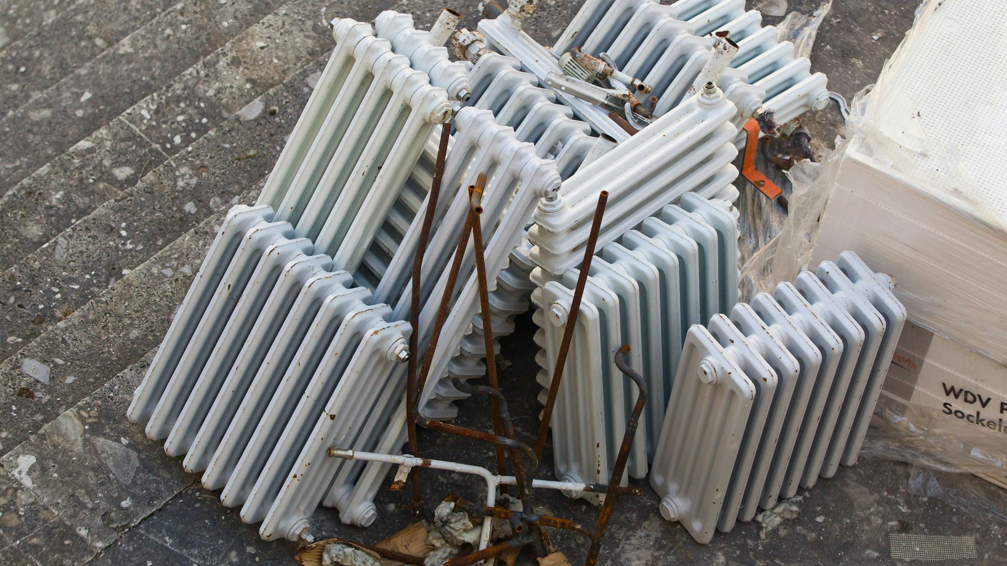 Alte ausrangierte Rippenheizkörper liegen nach einer Gebäude-Sanierung gestapelt in einem Innenhof. Daneben liegt Dämmmaterial zur Gebäudeisolierung.