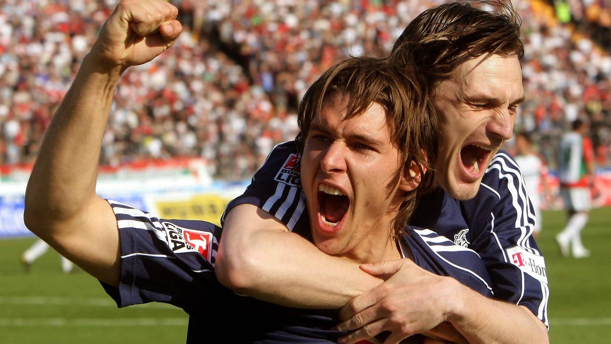 Ein Fußballer jubelt mit erhobenem Arm über seinen Torerfolg. Ein Mitspieler fällt ihm von hinten um den Hals.