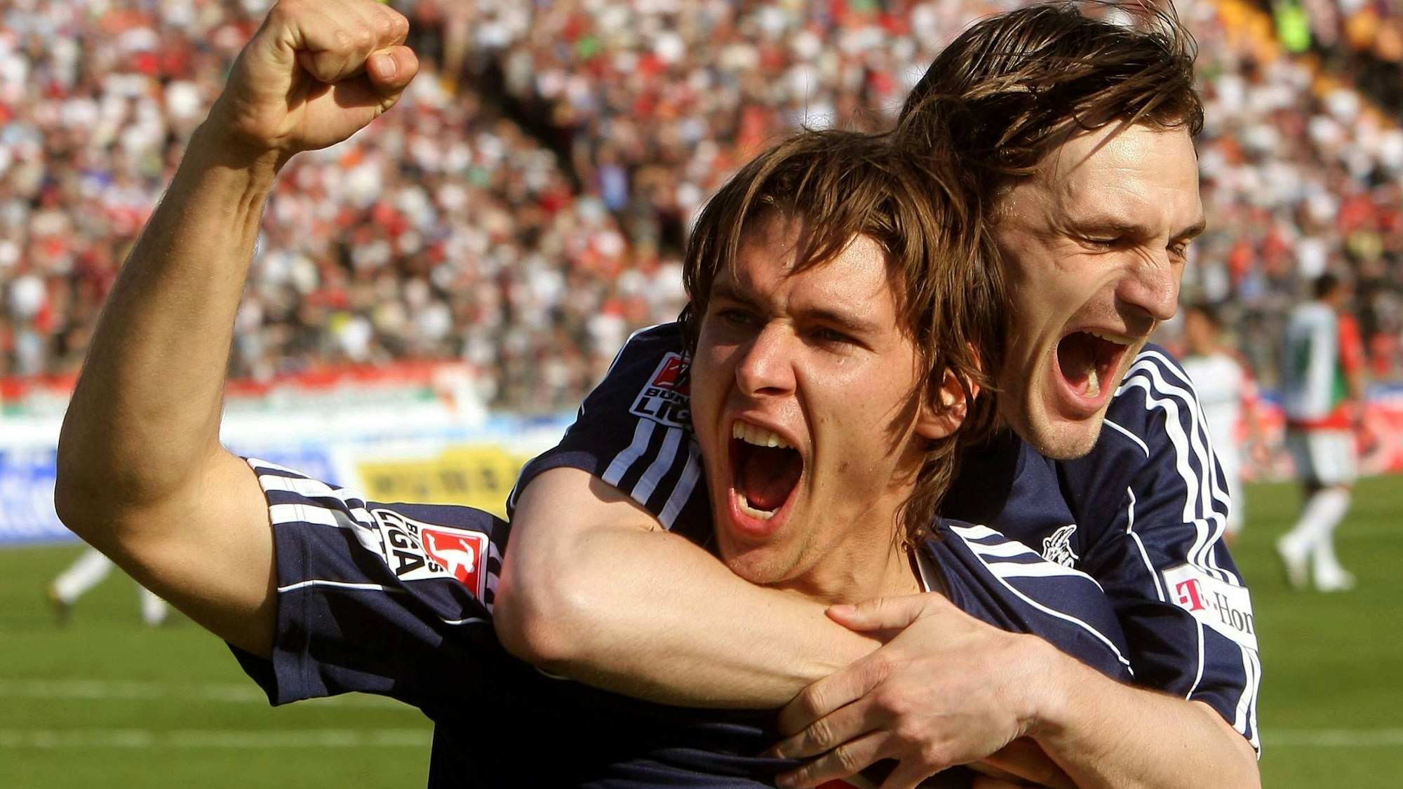 Ein Fußballer jubelt mit erhobenem Arm über seinen Torerfolg. Ein Mitspieler fällt ihm von hinten um den Hals.