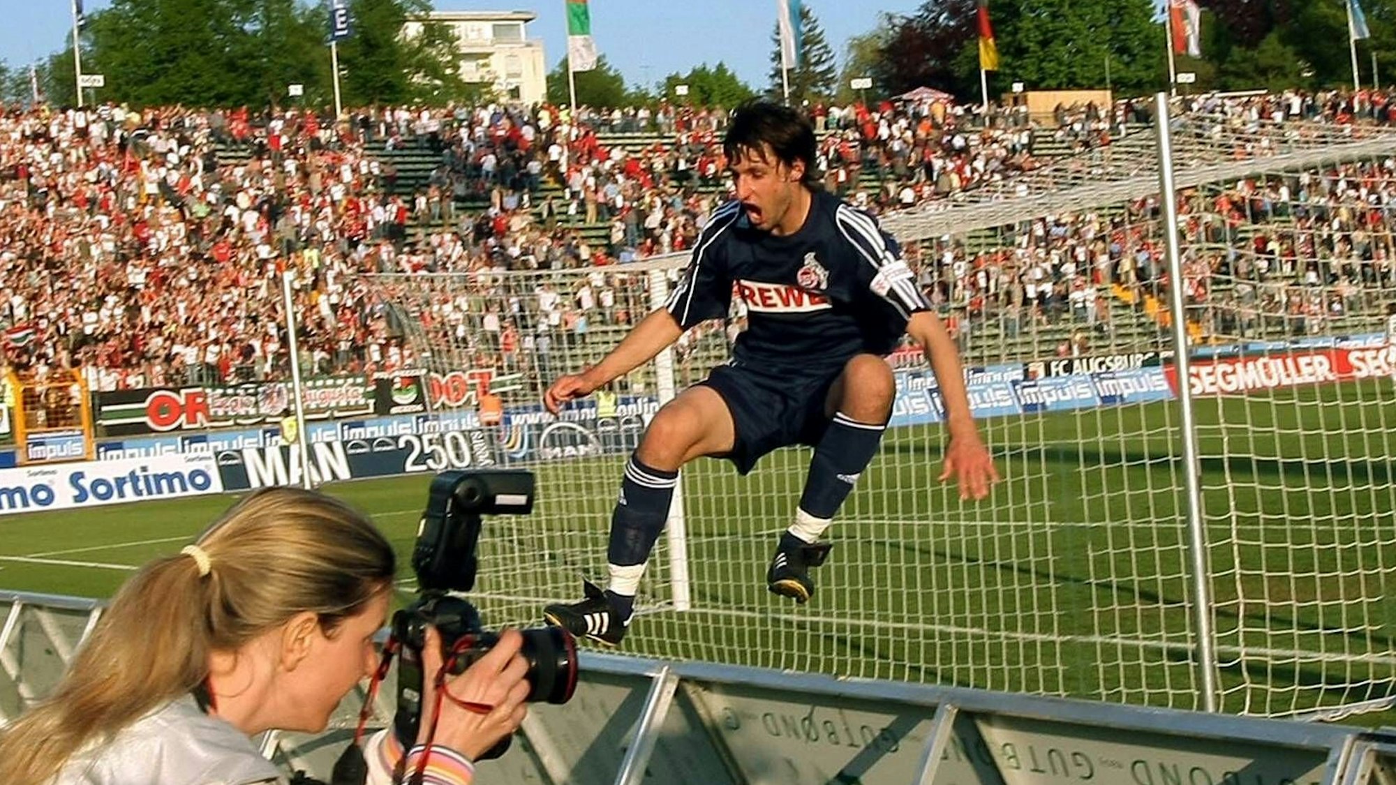 Ein Fußballer im dunkelblauen Trikot springt vor Freude über die Werbebande. Im Vordergrund ist eine Fotografin zu sehen.