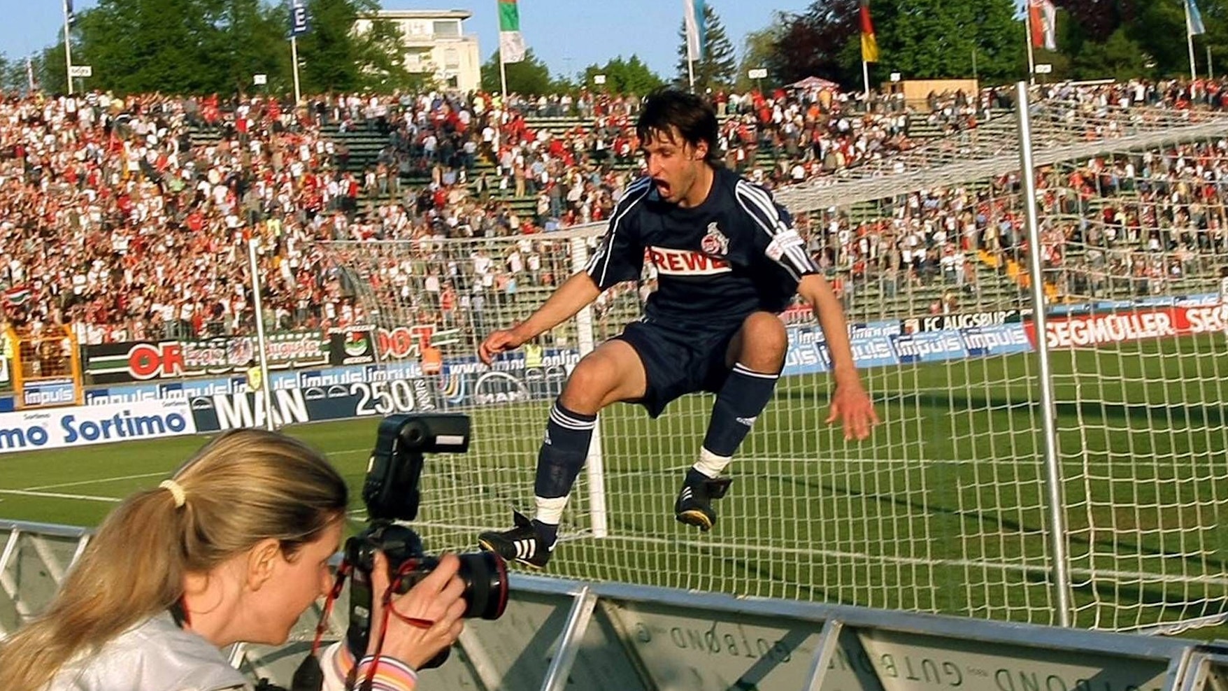 Ein Fußballer im dunkelblauen Trikot springt vor Freude über die Werbebande. Im Vordergrund ist eine Fotografin zu sehen.