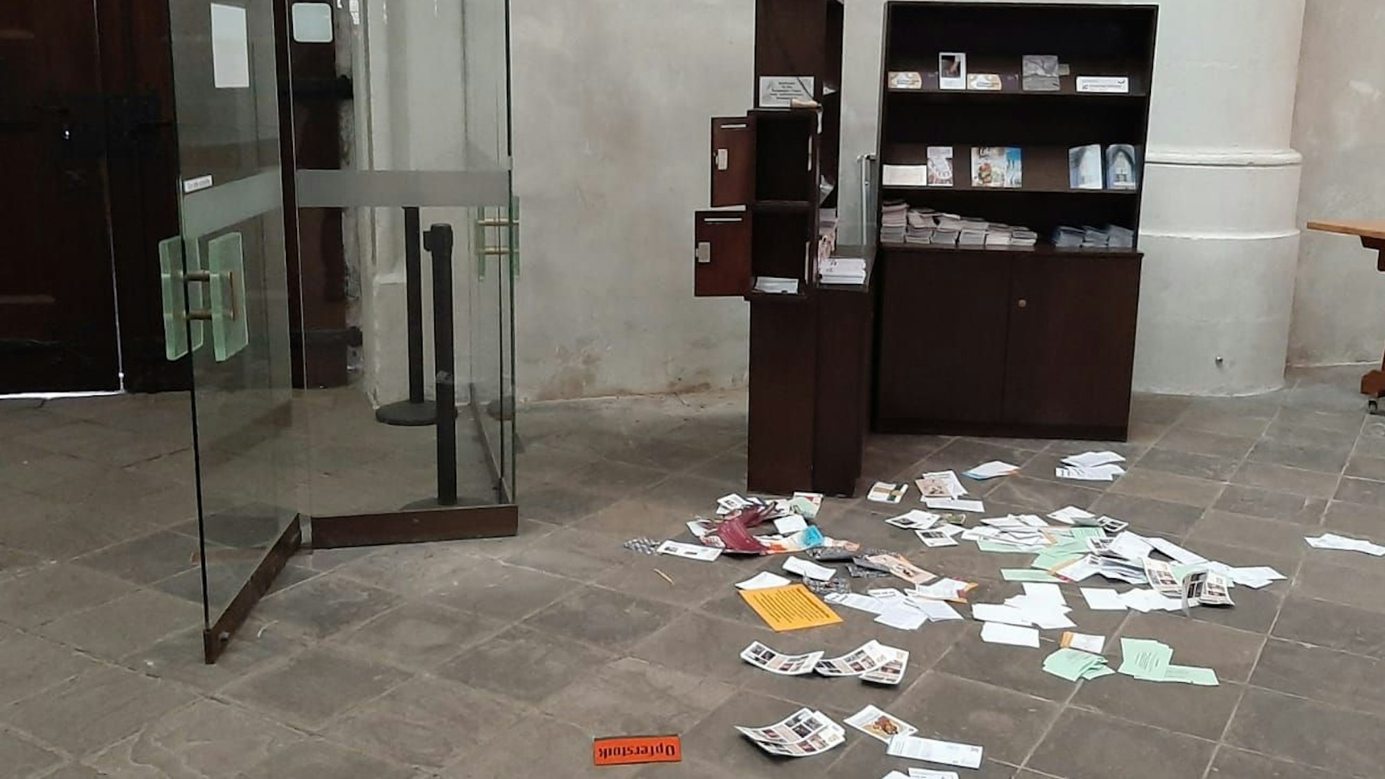 Broschüren und Schriften liegen auf dem Boden neben einer Kirchentür, der Schriftenstand ist aufgebrochen, Opferstöcke sind komplett gestohlen worden. Auf dem Boden liegt ein Schild mit der Aufschrift „Opferstock“.