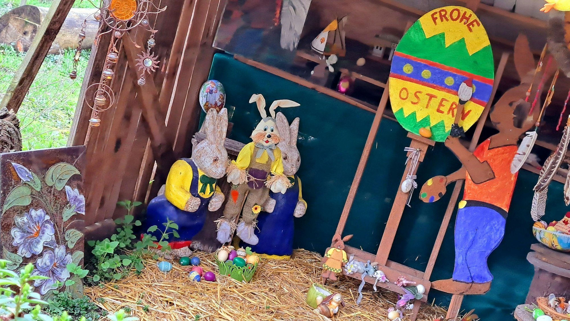 Hasenfiguren stehen in einem Unterstand, auf einem großen bunten Osterei steht „Frohe Ostern“.