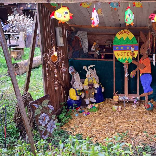 Hasenfiguren stehen in einem Unterstand, auf einem großen bunten Osterei steht „Frohe Ostern“.