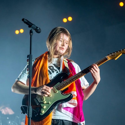 Girl in Red im Palladium. Sie steht am Stand-Mikrofon und spielt E-Gitarre. Um ihren Schultern liegt eine orange-weiß-pinke lesbische Pride-Flagge.