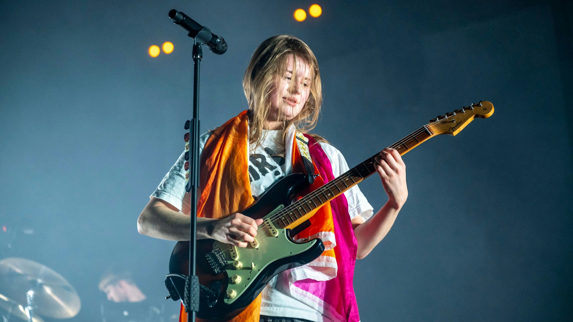 Girl in Red im Palladium. Sie steht am Stand-Mikrofon und spielt E-Gitarre. Um ihren Schultern liegt eine orange-weiß-pinke lesbische Pride-Flagge.