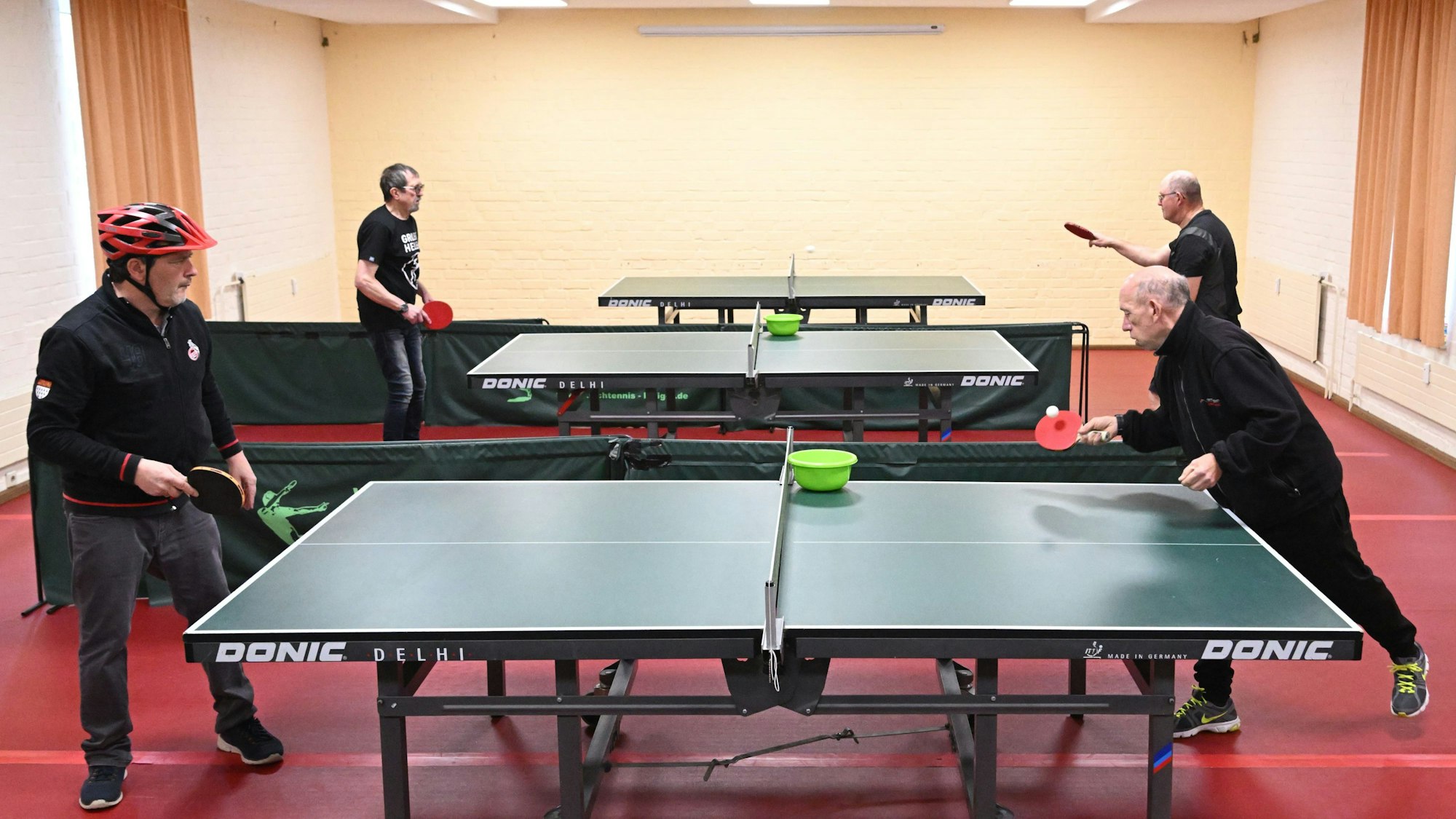 Drei Tischtennisplatten auf rotem Boden, vier ältere Herren in schwarzer Kleidung spielen an zwei Platten Tischtennis.