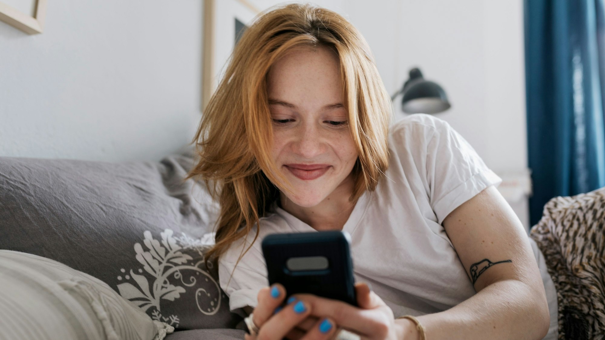 A woman smiling while lying on her bed at home and messaging on an online dating app using her smartphone.