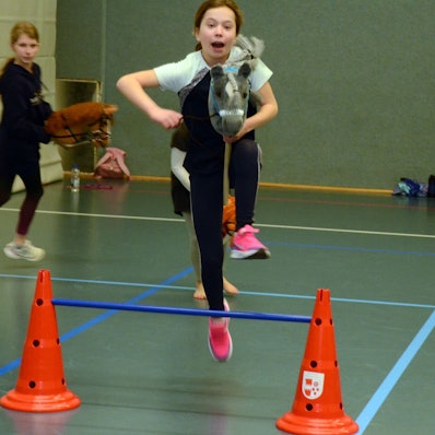 Es sind zwei Mädchen zu sehen die grade Sprünge mit ihren Steckenpferden üben.