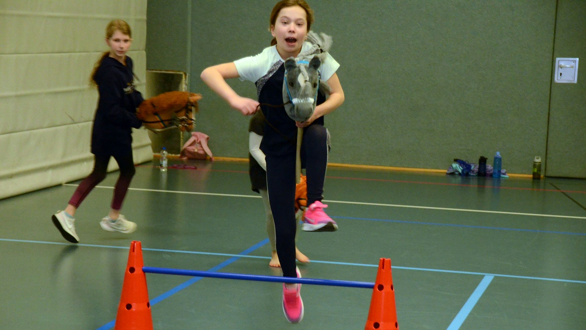 Es sind zwei Mädchen zu sehen die grade Sprünge mit ihren Steckenpferden üben.