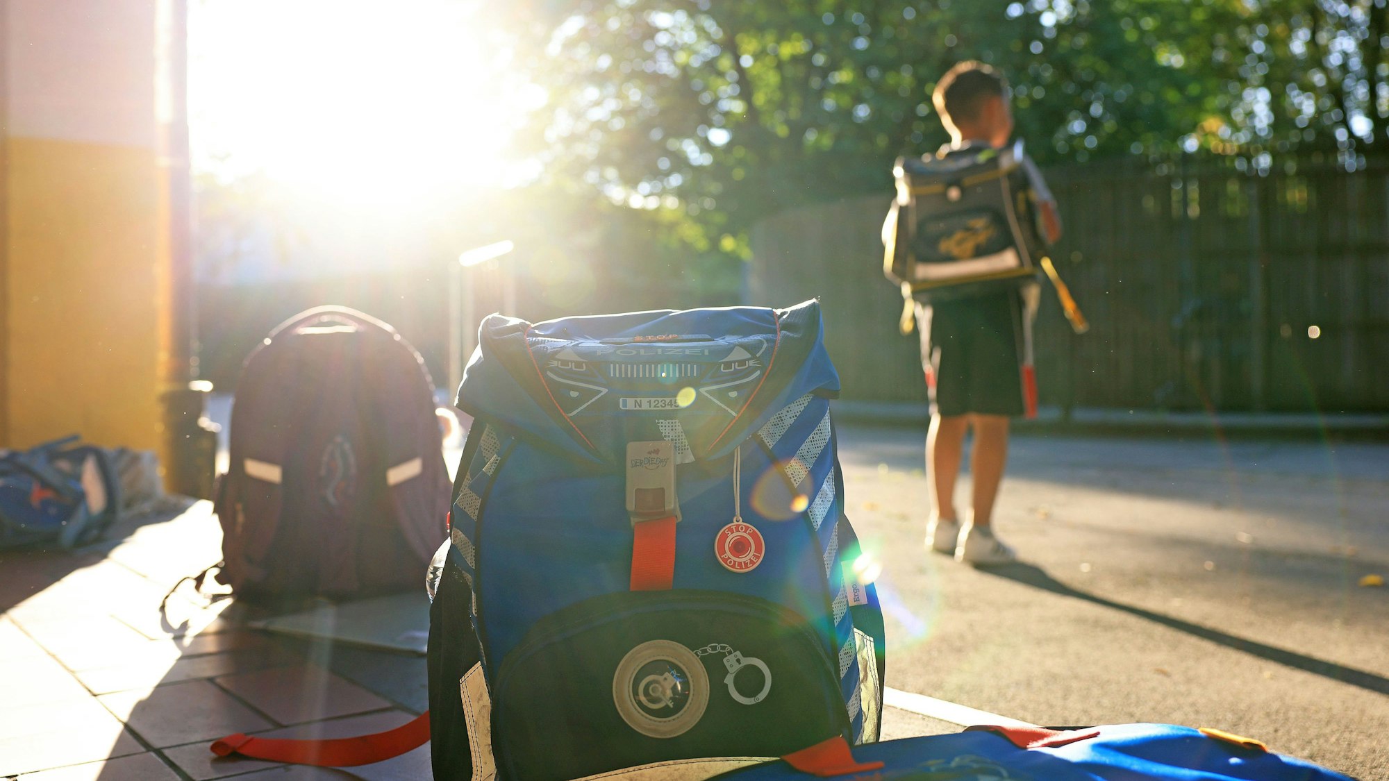 Schulranzen stehen am Morgen auf einem Schulhof. Ein Junge mit Schulranzen steht davor.