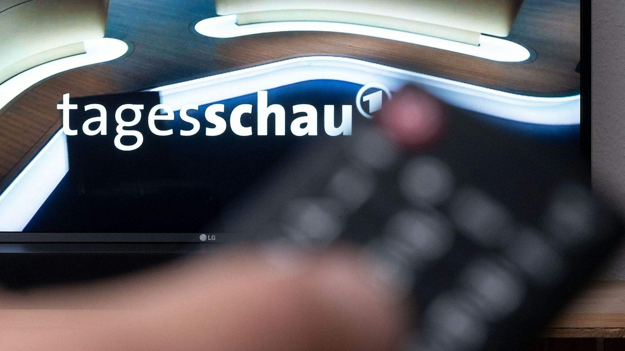 Ein Mann hält eine Fernbedienung vor einen Fernseher, auf dem die Tagesschau läuft.