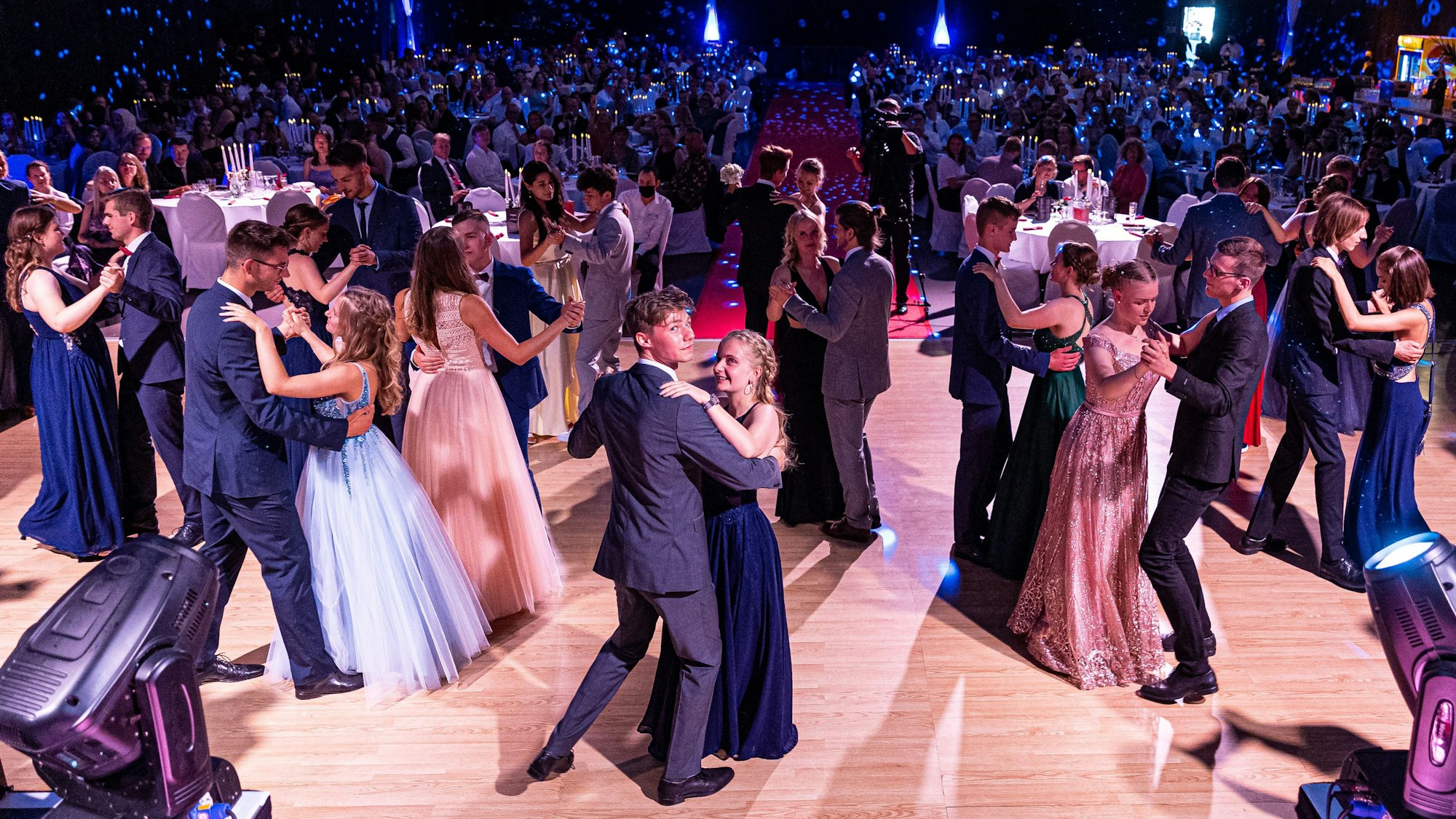 Im Bildvordergrund tanzen junge, in Abendkleider und Anzug gekleidete Menschen, während im Hintergrund Eltern an festlich dekorierten Tischen sitzen.