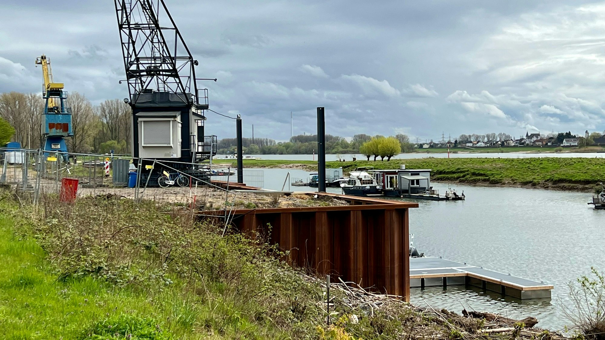 Hitdorfer Hafen, Kaimauer mit Krancafé im Vordergrund, hinten Hausboote im Hafen, dahinter der Rhein
