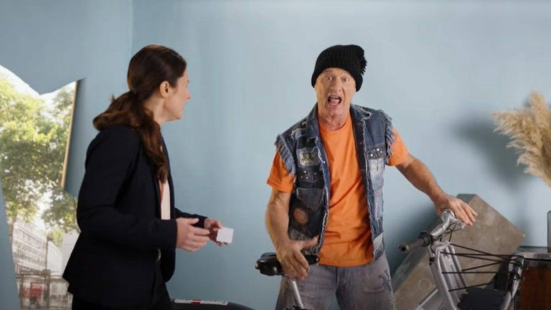 Tom Gerhardt steht mit KVB-Leihrad in einer Wohnung, neben ihm steht eine Frau im Anzug.