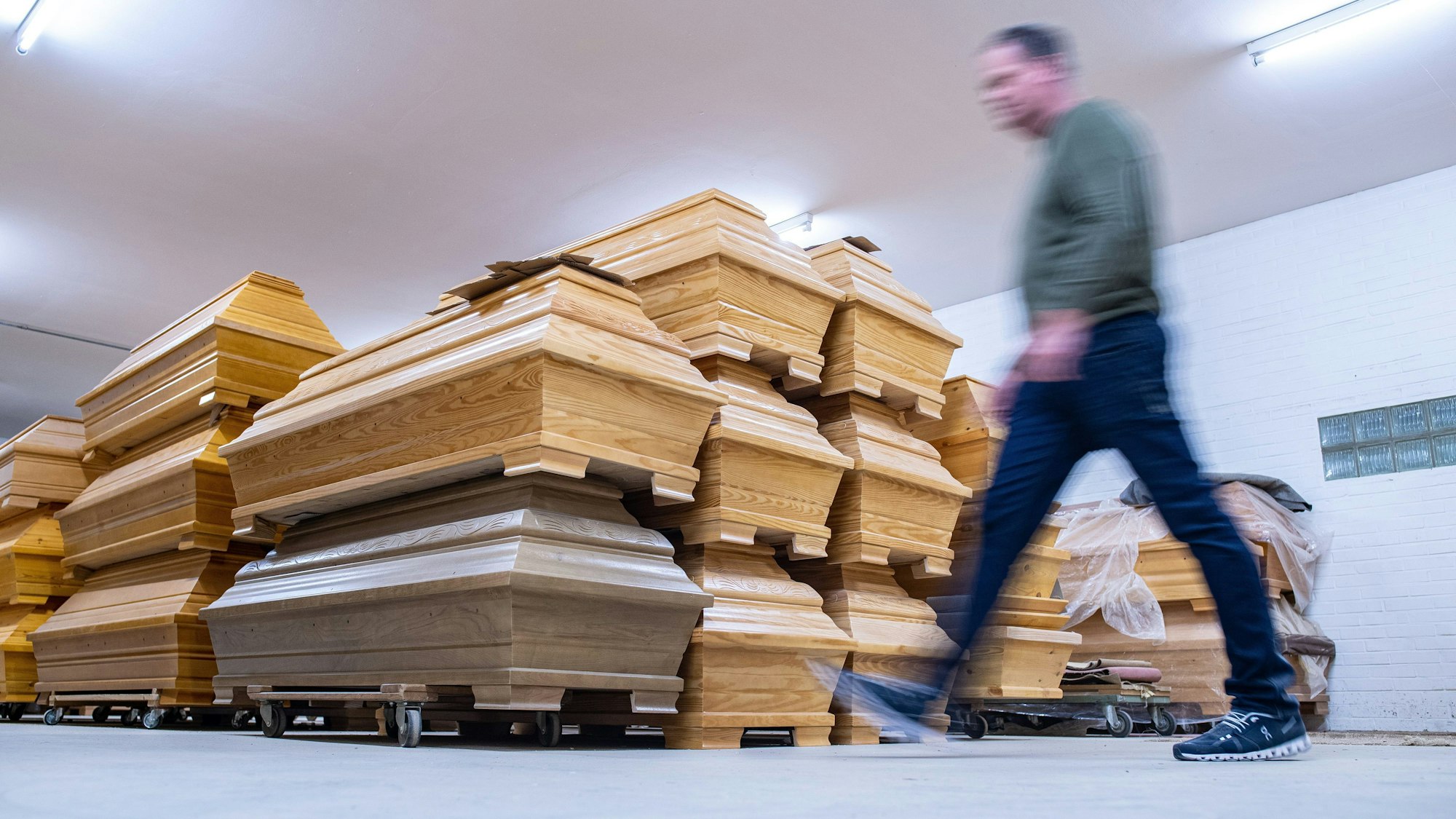Mehrere Särge aus Holz stehen übereinander gestapelt in einem Raum. Ein Mann geht an ihnen vorbei.