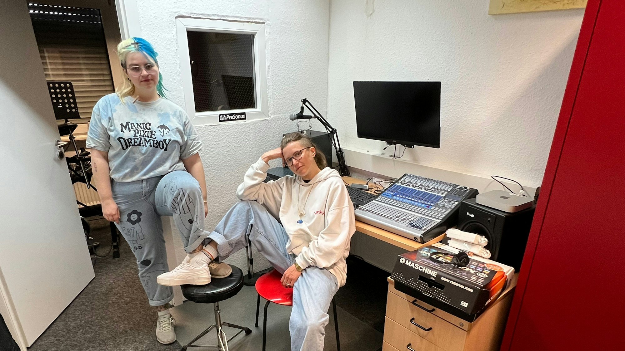 Zwei junge Frauen, sportlich gekleidet, sitzen lässig und mit jeweils einem Fuß auf einen Stuhl gestellt, in einem Tonstudio.