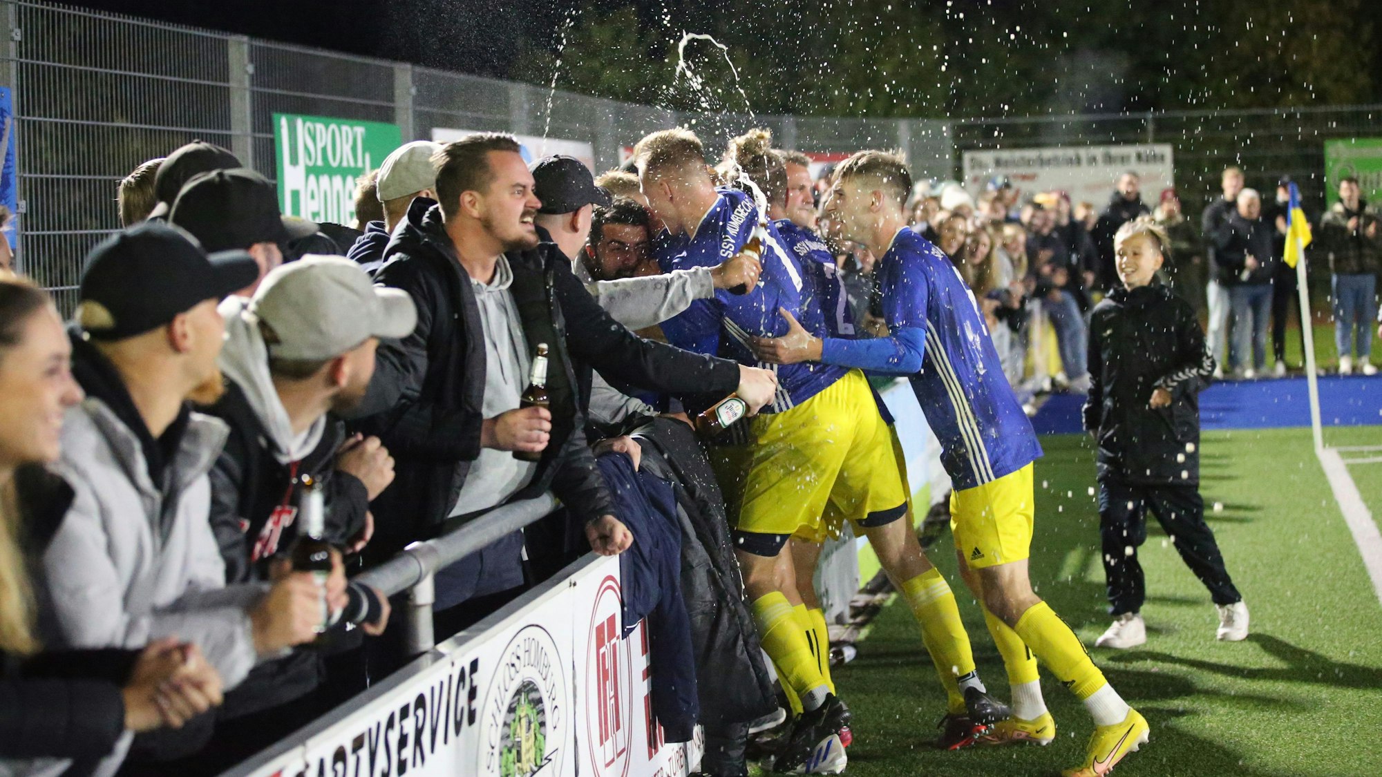 Fußballspieler jubeln mit ihren Fans, die hinter einer Bande am Spielfeldrand eines Fußballfeldes stehen.