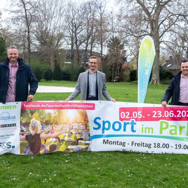 Die drei Männer posieren mit dem Banner von „Sport im Park“ und lächeln in die Kamera.
