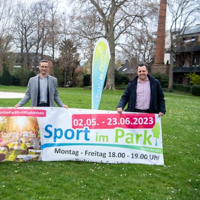 Die drei Männer posieren mit dem Banner von „Sport im Park“ und lächeln in die Kamera.
