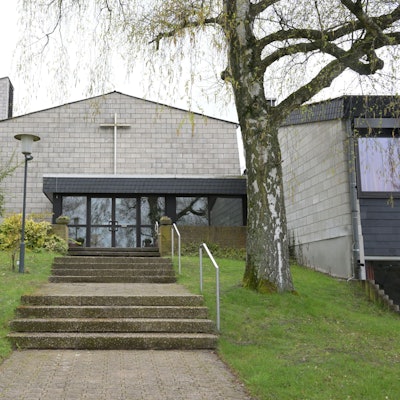 Vor dem Kirchengebäude mit Glockenturm und Kreuz über dem Eingang steht rechts ein Baum.