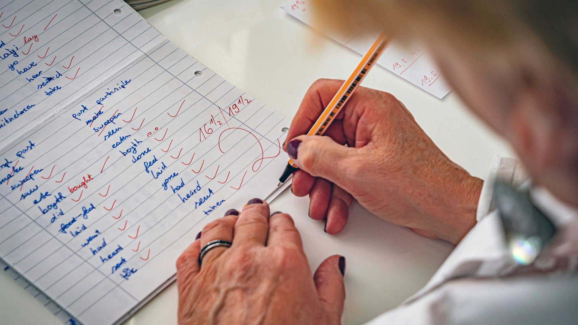 Auf einem DIN-A4-Blatt sind in zwei Spalten geschriebene Vokabeln mit Rotstift abgehakt. Die erreichte Punktzahl, die zu erreichende Punktzahl und die Note zwei sind ebenfalls rot vermerkt. Den Stift führt die rechte Hand einer Lehrerin, deren Kopf und Oberkörper am rechten Bildrand nur umrisshaft zu erkennen sind.