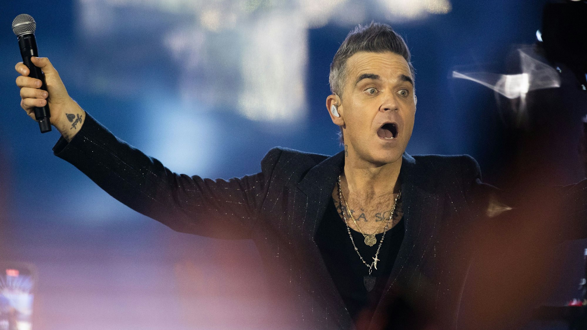 Robbie Williams singt auf der Bühne und reißt den Mund auf.