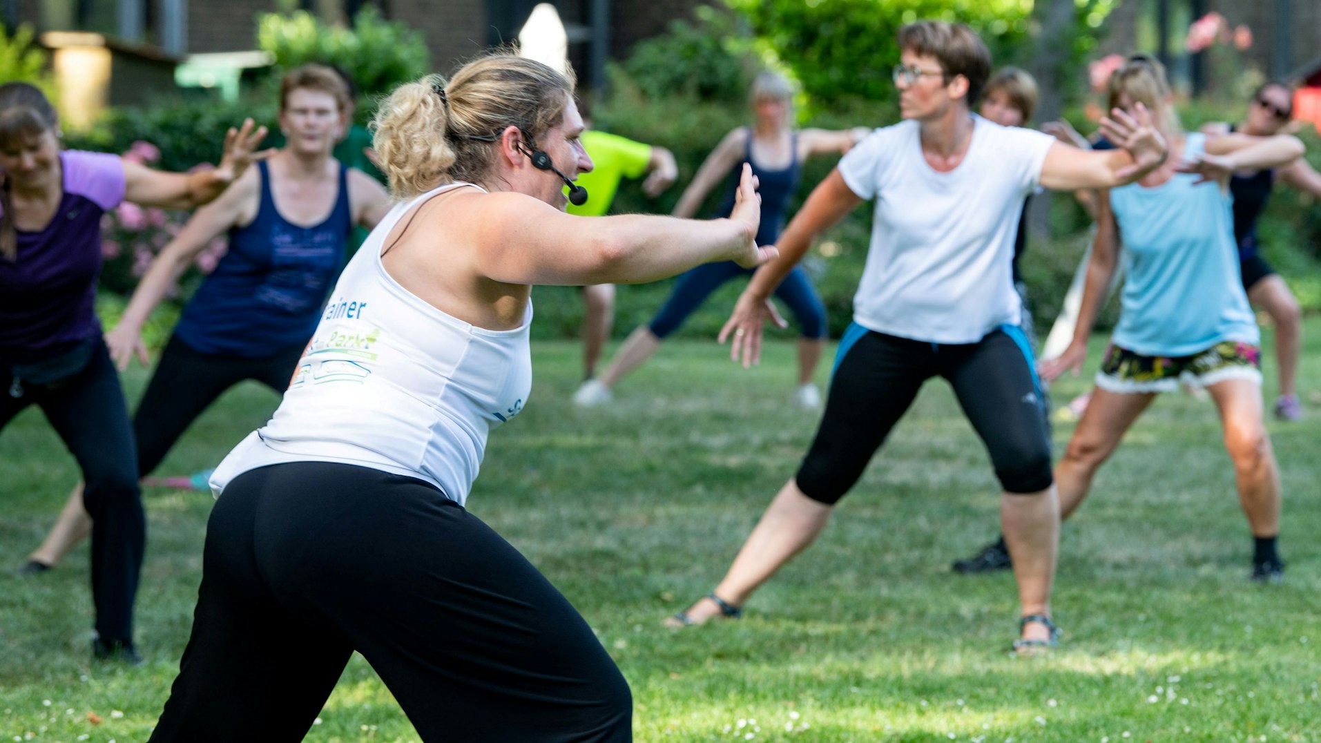 Aufnahme, wie Menschen bei einem Sportkurs mitmachen. Die Leiterin steht zu der Gruppe und trägt ein Mikrofon.