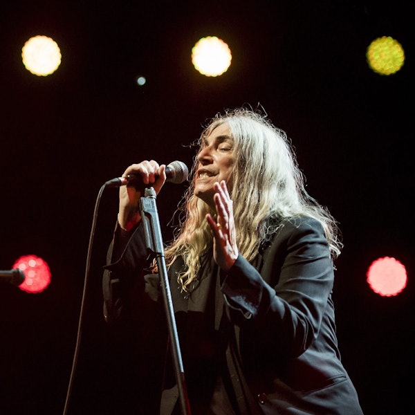 Die Sängerin hält ein Mikrofon nah an ihrem Mund. Sie trägt schwarze Kleidung und hat lange graue Haare.