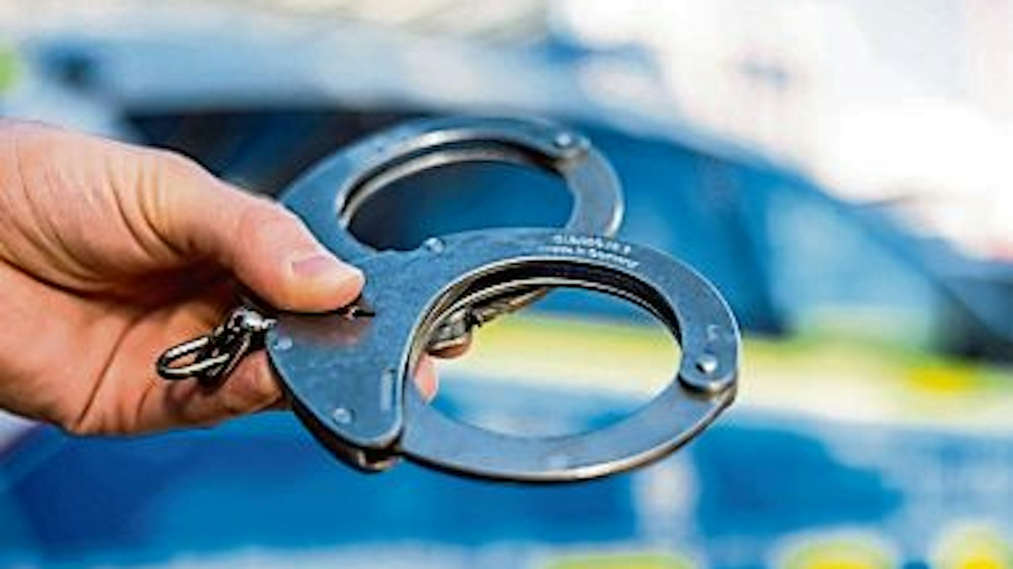 Das Foto zeigt Handschellen, im Hintergrund ist verschwommen ein Polizeiwagen zu sehen.