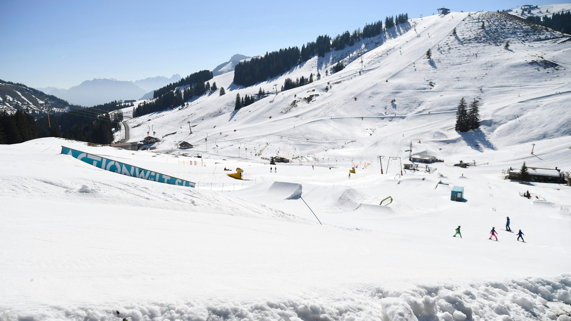Während sich im Vordergrund schon das erste Grün zeigt, fahren Skifahrer eine Piste am Sudelfeld im oberbayerischen Mangfallgebirge hinunter. (Symbolbild)