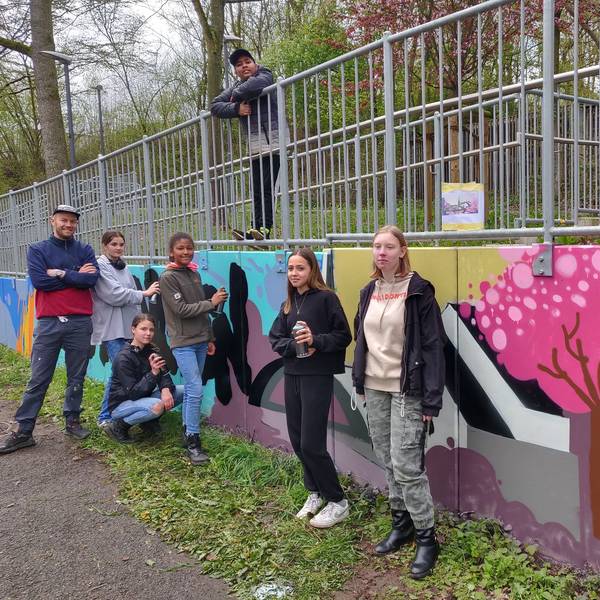 Die Graffiti-Künstler stehen mit dem ehrenamtlichen Workshop-Leiter Hannes Fries an der besprühten Mauer neben der Opladener Straße.